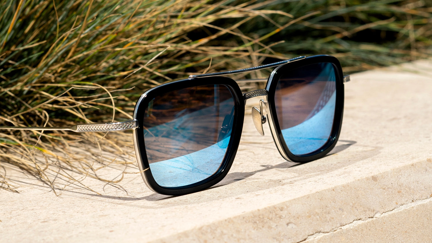 The image shows a stylish pair of square-shaped sunglasses with black frames and blue-tinted lenses, resting on a stone surface against a backdrop of grass.