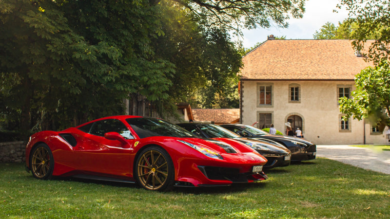 A red sports car parked on a lawn with several other luxury cars in the background, near a large, rustic building surrounded by trees.